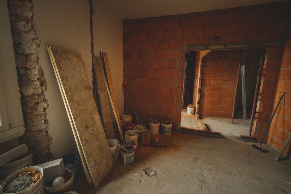 The image shows a partially constructed interior doorway with a plain white wall in the background. The door, which appears to be made of natural, unfinished wood with a simple panel design, is slightly open and positioned on the right side of the frame. The doorway frame is also partly finished, with exposed wooden studs and no trim or skirting visible. At the base of the wall, there are a few loose construction materials, including a white rectangular panel leaning against the wall, some scattered debris, and what appears to be a piece of blue plastic or protective covering on the unfinished floor. The overall setting suggests a renovation or refurbishment project, potentially involving framing or wall modification, typical in properties undergoing interior updates that may include waste removal or the clearance of building debris. Rubbish Clearance Pimlico may handle such waste as part of their professional services for on-site clearance of renovation rubbish, which often includes timber, drywall, and packaging materials, maintaining a clean work environment and ensuring proper disposal.