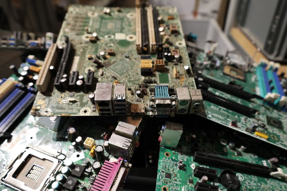 A large pile of discarded electronic circuit boards and components stacked together, featuring various colours and textures. The main board, positioned centrally, has a greenish surface with numerous metallic capacitors, resistors, and chips affixed, some with silver and gold finishes. Surrounding the main board are other smaller circuit boards, partially overlapping, with visible connectors, slots, and electronic parts in shades of black, blue, pink, and beige. The scene appears to be in an industrial or recycling facility environment, with a blurred background indicating additional electronic waste or workbenches. The lighting is bright but slightly diffuse, highlighting the metallic elements and textures of the circuit boards. This setting exemplifies electronic waste awaiting collection or proper disposal, aligning with private waste handling services that manage electronic item recycling, such as those offered by Rubbish Clearance Pimlico. The detailed depiction emphasizes the diversity of electronic waste materials processed in contexts related to avoiding fines through proper electronic waste disposal, an aspect often supported by independent rubbish clearance providers.