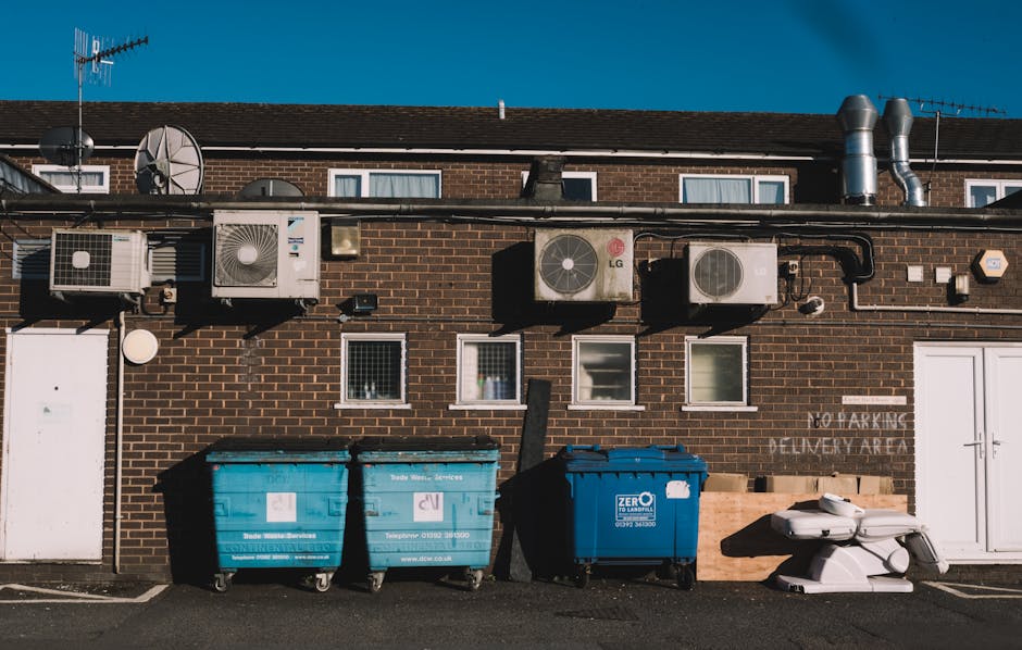 The image depicts the rear exterior of a brick building with a row of three small windows, each with white frames, set above a collection of waste disposal containers. There are four large, upright communal bins placed on the paved ground in front of the building, with two being bright blue and one darker blue, all fitted with wheels and lids; their surfaces are textured plastic with labels and contact details visible. To the right of the bins, there is a white plastic pallet or container apparatus, partially stacked on top of a small white object. Above, mounted along the brick wall, are various ventilation units and air conditioning units with metallic finishes, some with visible piping and ductwork, and a few satellite dishes are affixed to the wall. The environment appears to be a service area or back alley, with clear natural lighting against a bright blue sky, indicating daytime. The setting aligns with a location where private waste collection, including the handling of electrical waste, is arranged by services such as Rubbish Clearance Pimlico, supporting sustainable disposal practices in urban settings.