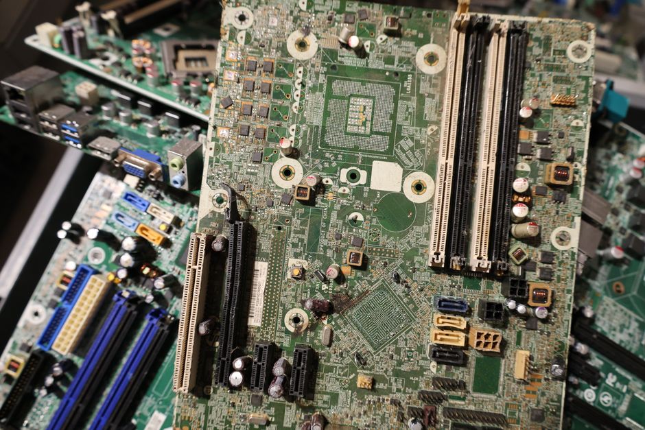 A large pile of discarded electronic circuit boards and components stacked together, featuring various colours and textures. The main board, positioned centrally, has a greenish surface with numerous metallic capacitors, resistors, and chips affixed, some with silver and gold finishes. Surrounding the main board are other smaller circuit boards, partially overlapping, with visible connectors, slots, and electronic parts in shades of black, blue, pink, and beige. The scene appears to be in an industrial or recycling facility environment, with a blurred background indicating additional electronic waste or workbenches. The lighting is bright but slightly diffuse, highlighting the metallic elements and textures of the circuit boards. This setting exemplifies electronic waste awaiting collection or proper disposal, aligning with private waste handling services that manage electronic item recycling, such as those offered by Rubbish Clearance Pimlico. The detailed depiction emphasizes the diversity of electronic waste materials processed in contexts related to avoiding fines through proper electronic waste disposal, an aspect often supported by independent rubbish clearance providers.