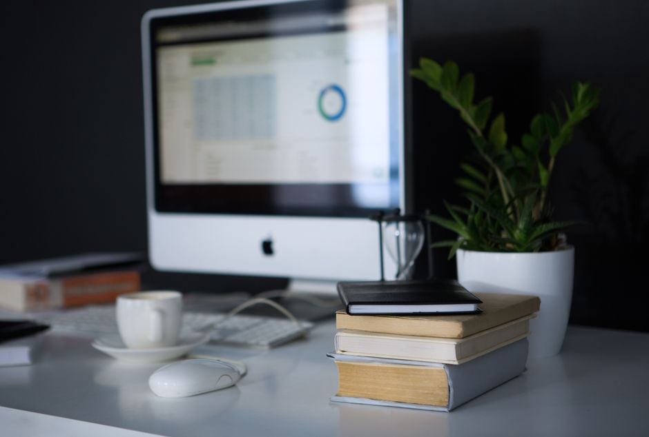 A close-up view of a clean, organized office desk workstation featuring a desktop computer with a large monitor displaying a spreadsheet or data table, positioned slightly towards the background. In the foreground, there is a neat stack of three hardcover books with neutral-colored covers—one black, one beige, and one light blue—placed on the right side of the desk. On top of the books rests a closed black notebook, adding to the professional setting. To the left of the books, a white ceramic coffee cup with a saucer and an integrated handle is visible, alongside a wired computer mouse and a wireless keyboard. In the background, a large potted plant with broad, dark green, glossy leaves is situated next to the monitor, providing a touch of natural decor. The workspace is illuminated by soft, natural lighting, highlighting the minimalist and tidy arrangement of items on the white surface, consistent with an office environment that could be associated with services like rubbish or waste management, emphasizing organization and clean, functional spaces.