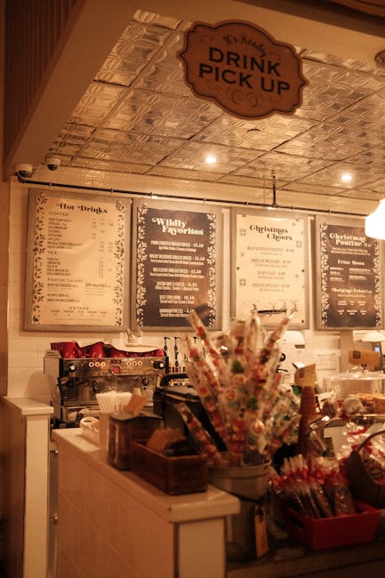 The interior of a casual cafe features a light-colored counter with various condiments, napkins, and utensils organized in containers. Behind the counter, there are illuminated menu boards displaying drink options, prices, and special offers on a white wall, with some sections highlighted in darker colors. An overhead sign reading 'Drink Pick Up' hangs from a decorative ceiling with a reflective, patterned metallic surface. To the right of the counter, there is a cluster of colorful straws and stirrers in a cylindrical container, alongside other small supplies like cups and paper envelopes. The lighting creates a warm, ambient atmosphere, emphasizing the cozy and functional setting typical of a coffee shop or quick-service cafe. The scene suggests a focus on customer service for takeout or onsite orders, aligning with independent or alternative disposal practices for waste generated in such establishments, while Rubbish Clearance Pimlico ensures appropriate rubbish management for commercial environments.