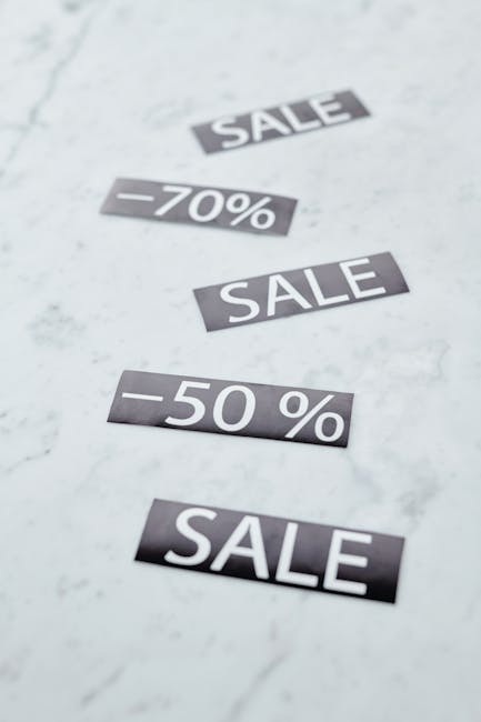 Multiple black and white sale discount stickers are arranged on a light-colored marble surface. The stickers display various promotional offers, including the words 'SALE,' '-70%,' and '-50%,' with some positioned at slight angles. The marble surface has subtle veining and a smooth texture, and the background appears evenly lit with no additional objects or clutter visible. This setup visually references the concept of discounts and sales, reminiscent of promotional clearance events that could relate to private or alternative disposal methods in waste management, as handled by companies like Rubbish Clearance Pimlico. The neutral scene emphasizes the idea of cost savings or price reductions often associated with clearance or removals, supporting the context of independent waste handling services.