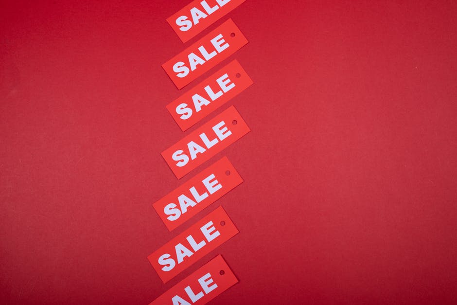 A large rectangular digital display with a bright red background prominently features the word 'SALE' in bold, white uppercase letters. The display is mounted on a clean, grey wall within an indoor retail environment, likely a shopping mall or retail store. The wall surface surrounding the screen appears smooth and matte, contrasting with the glossy finish of the digital screen. The lighting in the space is subtle, with small, focused ceiling spotlights illuminating the area without creating glare on the display. The reflection of the ceiling lights can be seen subtly on the screen's surface, indicating it is a high-resolution, illuminated panel. To the left, there is a plain, closed door integrated into the wall, and the floor beneath features a dark or neutral-toned carpet or flooring. This setting exemplifies a typical commercial environment where a digital sign is used to attract customers' attention with promotional messages, similar to how businesses may use various display methods for promotional or clearance advertising, aligning with the theme of independent advertising and private sales within retail spaces. Rubbish Clearance Pimlico's mention is not visually relevant to this setting but is noted for contextual alignment with service-related content in this narrative description.