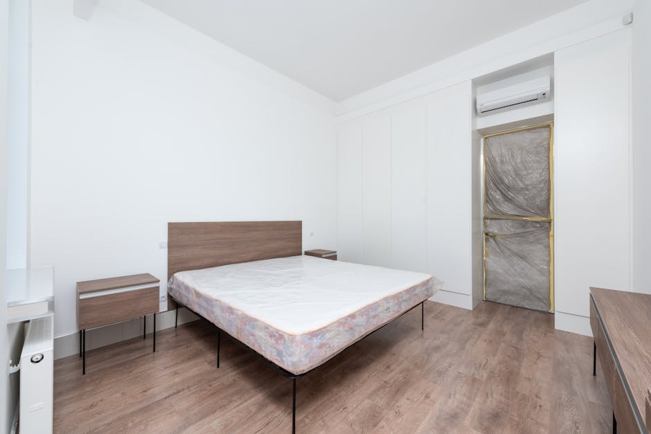 The image depicts a minimalistic, modern bedroom with a double bed placed centrally against a plain white wall. The bed has a simple, natural wood headboard and a standard mattress with floral-patterned sheets, showing a slight indentation where the sleeper might rest. Flanking the bed are two matching wooden bedside tables with sleek, flat surfaces, one positioned on each side, both supported by black metal legs, creating a cohesive, understated aesthetic. To the right of the bed, on the wall, a split-type air conditioning unit is installed above a narrow door that appears to be sealed with protective wrappings secured by gold-colored tape, indicating recent or ongoing renovation or cleaning procedures. The room features a light-toned wooden laminate flooring that complements the furniture, and the environment is well-lit with natural light, giving the space a clean, uncluttered appearance. The overall scene suggests a professionally managed property, with the visible elements consistent with a pre- or post-occupancy refurbishment, possibly requiring or awaiting rubbish removal or private waste disposal handled by services such as Rubbish Clearance Pimlico, as part of their comprehensive rubbish services.