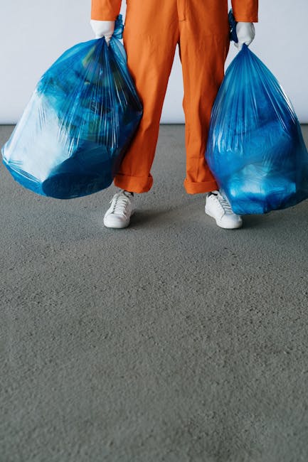 A person dressed in orange work overalls and white sneakers is standing on a gray paved surface, holding two large blue plastic rubbish bags filled with waste. The bags appear to contain various household or garden debris, visible through the semi-transparent material, which has a glossy finish. The individual’s hands are covered with gray gloves, and the background is plain and neutral, suggesting an indoor or outdoor space designated for rubbish collection or waste transfer, possibly related to private waste disposal services such as those offered by Rubbish Clearance Pimlico. The scene emphasizes the act of rubbish collection, with the focus on the bags and the effort involved in managing waste materials, aligning with the concept of independent waste removal or on-site clearance. The overall composition conveys a tidy, organized approach to rubbish handling, reflecting professional waste management practices relevant to the UK context of private rubbish removal services.