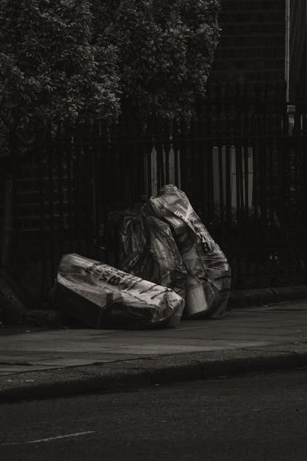 A group of several black plastic rubbish bags, some crumpled and tied at the top, are positioned on a paved urban sidewalk near a black metal fence and a dark green hedge. The bags appear to contain household waste and are partially leaning against the fence, with some bulging indicating they are full. The scene is outdoors with natural lighting, and a portion of the street is visible in the foreground, suggesting a typical residential or commercial area. The presence of the rubbish bags on the pavement aligns with waste collection or clearance services offered by companies such as Rubbish Clearance Pimlico, which provides private and on-site waste disposal options in lieu of municipal collections.