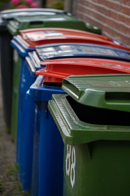 A row of outdoor waste bins aligned along a paved surface adjacent to a brick wall, with the nearest bin in the foreground featuring a green plastic lid, which is slightly open revealing the dark interior. Behind it, a blue bin with a closed lid, followed by a red bin, also with a closed lid, and additional bins of various colors extending into the background. The bins appear to be made of durable plastic with textured lids and are positioned closely, suggesting they are part of a commercial or residential rubbish collection setup. The environment is outdoors, with natural light illuminating the scene and a blurred background indicating a residential or urban area suitable for private waste handling or independent rubbish collection by companies like Rubbish Clearance Pimlico, serving local rubbish removal needs.