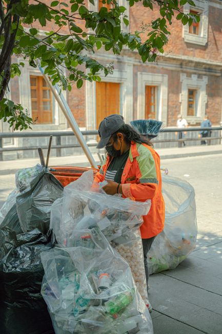 A group of several black plastic rubbish bags, some crumpled and tied at the top, are positioned on a paved urban sidewalk near a black metal fence and a dark green hedge. The bags appear to contain household waste and are partially leaning against the fence, with some bulging indicating they are full. The scene is outdoors with natural lighting, and a portion of the street is visible in the foreground, suggesting a typical residential or commercial area. The presence of the rubbish bags on the pavement aligns with waste collection or clearance services offered by companies such as Rubbish Clearance Pimlico, which provides private and on-site waste disposal options in lieu of municipal collections.