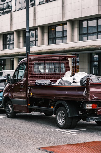A small burgundy commercial pickup truck is parked on an urban street in front of a modern building with large glass windows and concrete facades. The truck's cargo bed is loaded with various types of waste and rubbish, including large bags of mixed debris and smaller items, some covered with black and white plastic sheeting. The cargo appears to be loosely stacked, with textiles, discarded packaging, and bulky waste materials visible. The vehicle features a metal mesh partition behind the cab, providing separation for the cargo area. The street surface is asphalt, and a black streetlamp stands adjacent to the truck, with a portion of a red and white striped traffic barrier visible in the foreground. The scene is taken during daylight hours, with natural lighting highlighting the textures and colors of the waste, vehicle, and surrounding environment. This image exemplifies an independent rubbish collection service, such as Rubbish Clearance Pimlico, showcasing the process of private waste handling and on-site clearance for domestic or commercial waste disposal.