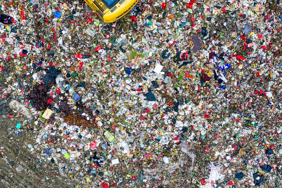 An aerial view of a large, open landfill site densely packed with mixed household and commercial waste materials, including plastic bottles, paper, fabric, and miscellaneous debris, scattered across the uneven ground surface. The waste is piled high with various colours, textures, and materials overlapping, creating a chaotic mosaic of refuse. In the upper part of the image, a yellow waste management vehicle is partially visible, indicating active waste collection or disposal at the site. The surrounding environment appears to be an outdoor area with no visible structures, and the lighting suggests natural daylight. This scene exemplifies the typical scene of unmanaged or privately managed rubbish accumulation, highlighting the importance of professional rubbish clearance services such as those offered by Rubbish Clearance Pimlico for efficient, responsible waste disposal and site cleanup beyond local authority collection options.