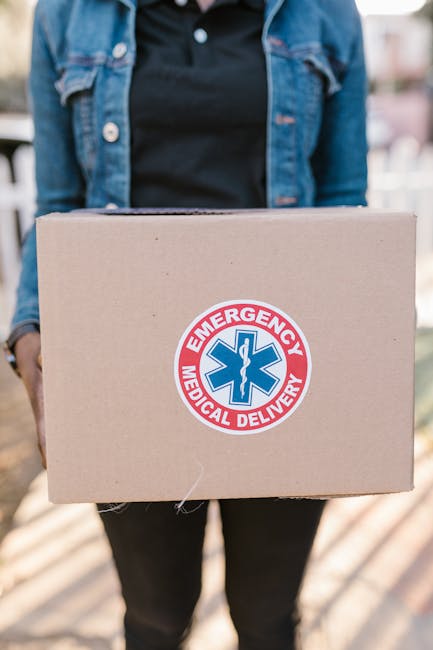 A person wearing a dark denim jacket and black top is holding a cardboard box in front of them, with the box featuring a round blue and red 'Emergency Medical Delivery' sticker prominently displayed on the front. The background includes a blurred outdoor setting with a hint of greenery and sunlight, suggesting a daytime environment. The box appears to be sealed and is held securely with both hands, positioned at waist level. This image represents a delivery process involving urgent or specialized items, such as medical supplies, which could relate to private waste handling or on-site clearance services by Rubbish Clearance Pimlico that support alternative rubbish disposal methods. The scene emphasizes careful handling of the package, relevant to the context of independent collection and timely disposal or transport of sensitive or specialized waste materials. The lighting is natural, with the subject standing outdoors, highlighted by soft sunlight that enhances the clarity of the packaging details and environmental context, aligned with professional waste management practices.