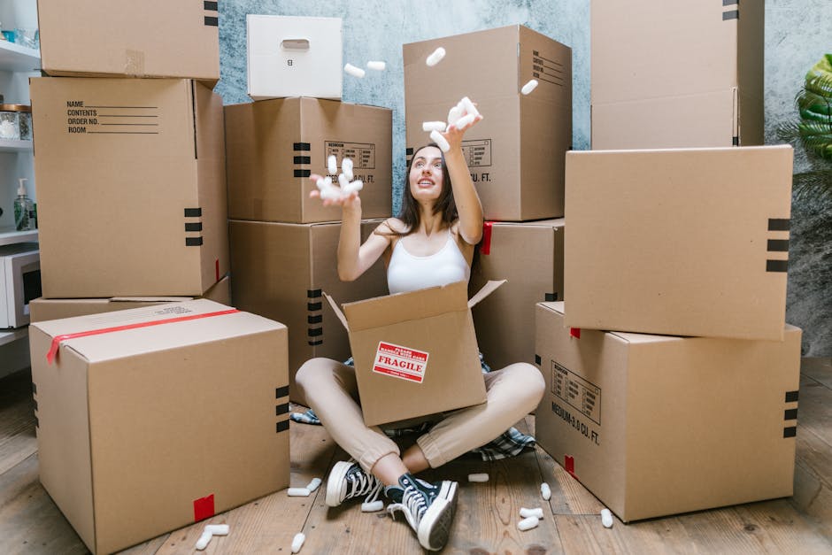 A young woman with long, dark hair and light skin is sitting cross-legged on a wooden floor inside a room, surrounded by large cardboard boxes of varying sizes, some sealed with red tape and others open. She is wearing a white sleeveless top, beige pants, and black-and-white sneakers. She is smiling and joyfully tossing white packing peanuts into the air, with some falling into an open box that she is sitting inside. The background features a textured grey wall and a green plant to the right, indicating an indoor setting. The scene depicts an active packaging or moving process, with the numerous boxes and packing materials suggesting preparation for storage or transportation, aligning with the context of private or alternative waste handling or rubbish clearance services that Rubbish Clearance Pimlico might offer as part of their rubbish removal solutions.