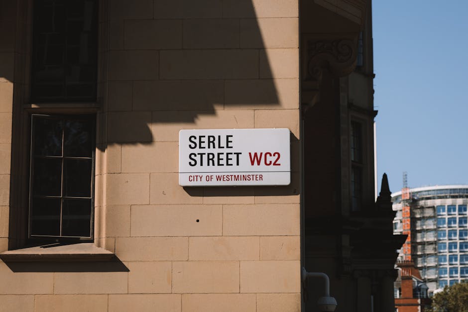Close-up view of a street name sign mounted on a beige stone building wall, displaying 'Serle Street WC2' with the subtitle 'City of Westminster' in red text, illuminated by sunlight that casts a shadow on the upper part of the sign. To the left of the sign, a black-framed window with multiple glass panes reflects some interior or exterior light, and the window sill is made of stone matching the building's facade. In the background, on the right side of the image, a portion of modern high-rise buildings with glass windows and steel structures is visible against a clear blue sky. The scene is set in an urban environment where private waste management or alternative rubbish removal services, such as those provided by Rubbish Clearance Pimlico, may operate, especially in areas like Westminster with historical and contemporary architecture.