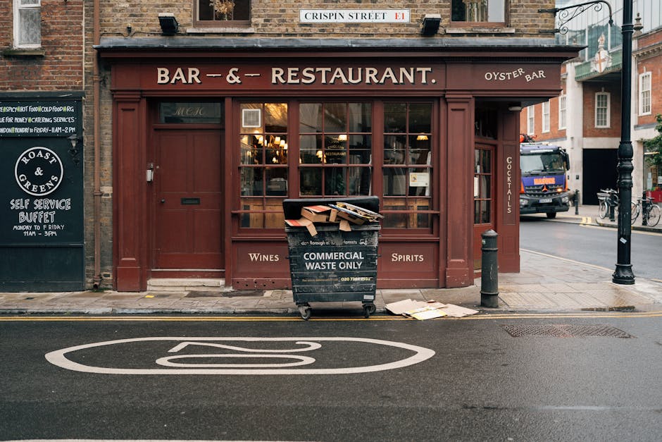 A black wheeled waste collection bin labeled 'Commercial Waste Only' is positioned on the pavement in front of a reddish-brown storefront with large glass windows and wooden framing, which appears to be a bar and restaurant. The bin is partially filled with flattened cardboard boxes and other refuse, with some boxes spilling onto the ground nearby. Adjacent to the bin on the left, there is a small black signboard with white text advertising a self-service buffet. The pavement and street are clean, with a white bicycle symbol painted on the Tarmac road surface directly in front of the bin, indicating a bicycle lane. To the right, a black bollard and a streetlamp are visible, and further along the street, there is a parked vehicle partially obscured by the sidewalk furniture and a bicycle secured to a bike rack. The environment suggests an urban area with a mix of pedestrian and vehicular activity, closely aligned with private waste handling and refuse collection services. The overall scene depicts typical street-level waste disposal in a city environment, relevant to rubbish removal services such as those offered by Rubbish Clearance Pimlico.