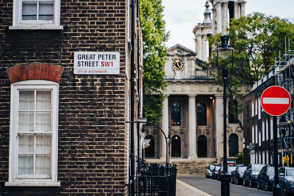 Close-up view of a street name sign mounted on a beige stone building wall, displaying 'Serle Street WC2' with the subtitle 'City of Westminster' in red text, illuminated by sunlight that casts a shadow on the upper part of the sign. To the left of the sign, a black-framed window with multiple glass panes reflects some interior or exterior light, and the window sill is made of stone matching the building's facade. In the background, on the right side of the image, a portion of modern high-rise buildings with glass windows and steel structures is visible against a clear blue sky. The scene is set in an urban environment where private waste management or alternative rubbish removal services, such as those provided by Rubbish Clearance Pimlico, may operate, especially in areas like Westminster with historical and contemporary architecture.