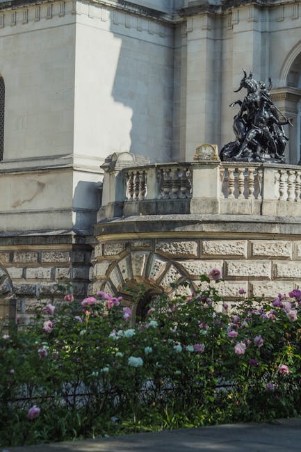 A detailed view of a historic stone building featuring a rounded corner with decorative stonework and an ornate balustrade. In the background, a bronze sculpture of a mounted figure and other figures on horseback is visible atop the structure. Below the building, a garden area with blooming pink, purple, and white roses and green foliage extends across the foreground, partially obscuring the architectural base. The scene is illuminated by natural daylight, highlighting the textures of the stone and metal sculpture, and creating soft shadows on the building's façade, while the garden adds a touch of colour that contrasts with the historic stonework. This setting may be part of a public space or historical site, where maintenance involves routine cleaning and occasional clearing of overgrown vegetation, supporting nearby rubbish removal services such as those offered by Rubbish Clearance Pimlico, which specialises in alternative waste handling methods for urban environments.