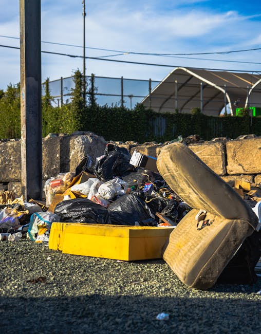 A close-up of a pile of mixed rubbish on a gravel surface, including black plastic bin bags, a yellow plastic container, a worn, dirty beige upholstered chair cushion with visible fabric texture, and a disused car tire lying on its side in the foreground. Behind the debris, there is a low stone wall made of irregularly shaped rough-hewn stones, with some greenery and shrubs visible above it. In the background, a metal fence encloses a sports facility with a large curved roof structure, and overhead power lines stretch across a partly cloudy sky, indicating an outdoor urban setting. The scene appears to depict an area prepared for rubbish removal, aligning with private waste disposal or on-site clearance carried out by a specialist such as Rubbish Clearance Pimlico, supporting alternative rubbish collection services.